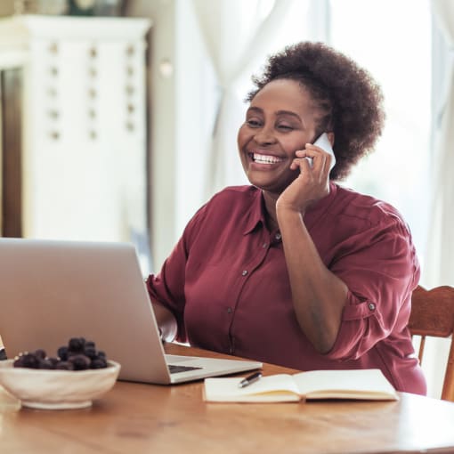 A person sits at a table with a laptop and a coffee cup and talks to a colleague.